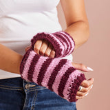 A person wearing pink and burgundy striped knit fingerless wrist warmers, paired with a white t-shirt and blue jeans, against a solid dusty pink background.