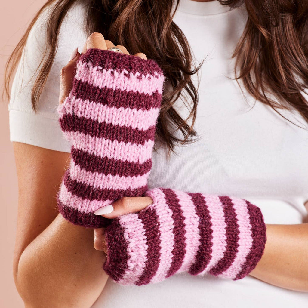 A close-up of a person wearing pink and burgundy striped knit fingerless wrist warmers, holding one up to show the texture and pattern against a soft pink background.