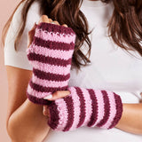 A close-up of a person wearing pink and burgundy striped knit fingerless wrist warmers, holding one up to show the texture and pattern against a soft pink background.