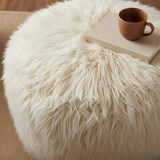A close-up of a fluffy faux fur rug in a warm cream or light beige color, styled with a book and a cup in the corner.