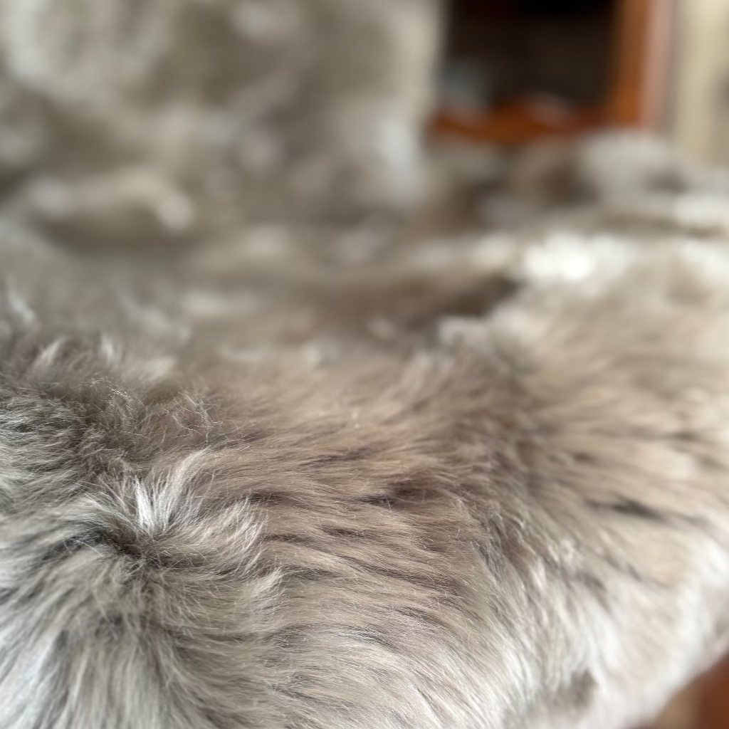 Close-up of a shaggy faux fur surface in soft gray and beige tones, with a blurred indoor background.