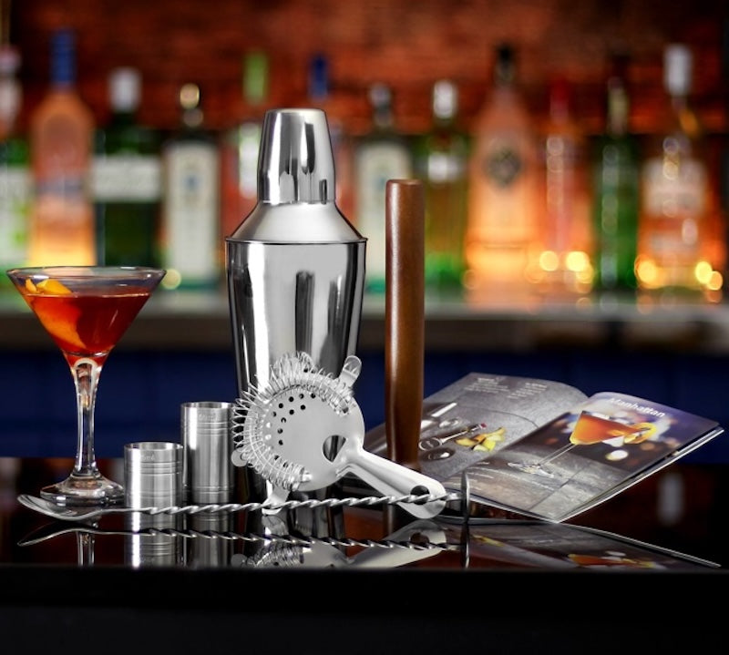 A stainless steel cocktail shaker set on a bar counter, including a shaker, strainer, jiggers, and bar tools, with a filled martini glass and an open magazine in front of a blurred bar background with liquor bottles.