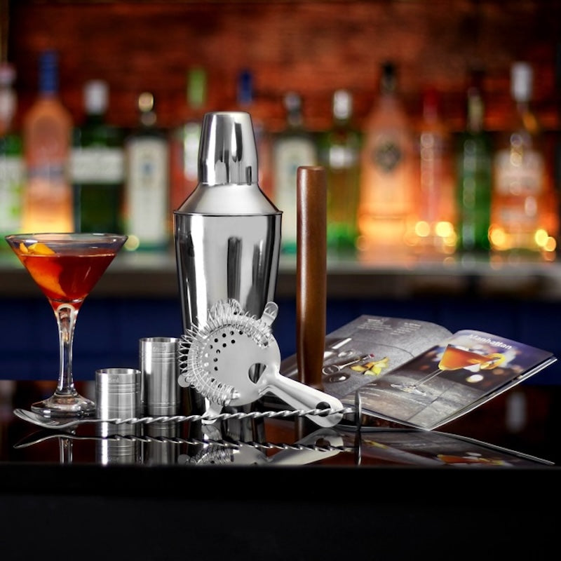 A stainless steel cocktail shaker set on a bar counter, including a shaker, strainer, jiggers, and bar tools, with a filled martini glass and an open magazine in front of a blurred bar background with liquor bottles.