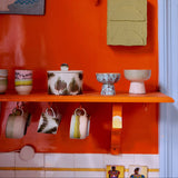 Orange wall with shelves displaying ceramic items and hanging mugs.