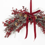 A festive Christmas wreath made of gray twig branches, covered with clusters of red berries and small pinecones, tied with a wide red velvet ribbon bow at the top against a plain white background.