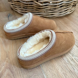 A pair of tan suede slip-on slippers with a soft cream sherpa lining and a braided trim around the opening, displayed on a light wood floor with a wicker basket in the background.