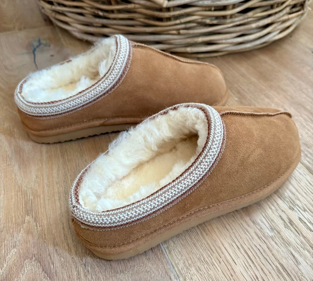 A pair of tan suede slip-on slippers with a soft cream sherpa lining and a braided trim around the opening, displayed on a light wood floor with a wicker basket in the background.