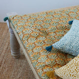 A close-up of a patterned upholstered bench or daybed with a mustard yellow and teal leafy print, styled with two decorative throw pillows on a woven rug.