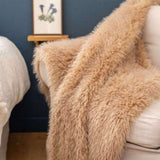 A fluffy, long-pile sheepskin or faux-sheepskin throw blanket in a warm camel beige color, draped over a cream-colored armchair in a cozy room setting with a dark blue wall and a small wooden shelf in the background.