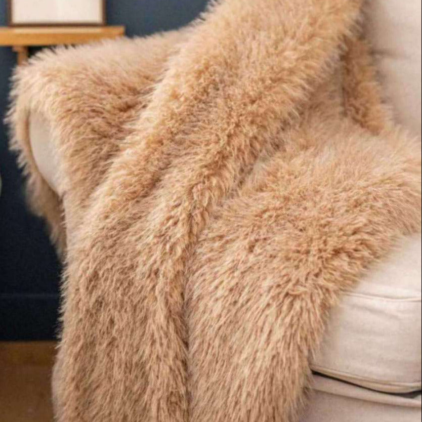 A fluffy, long-pile sheepskin or faux-sheepskin throw blanket in a warm camel beige color, draped over a cream-colored armchair in a cozy room setting with a dark blue wall and a small wooden shelf in the background.