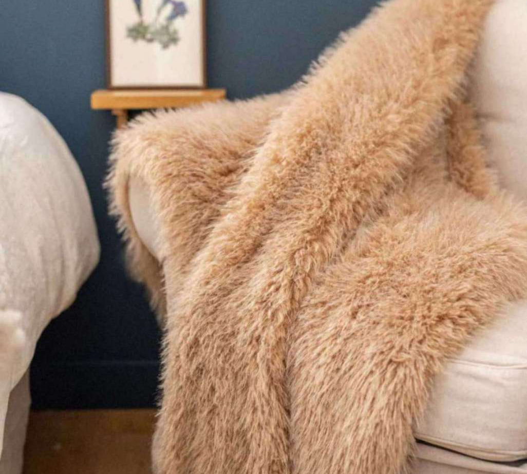 A fluffy, long-pile sheepskin or faux-sheepskin throw blanket in a warm camel beige color, draped over a cream-colored armchair in a cozy room setting with a dark blue wall and a small wooden shelf in the background.