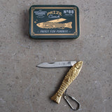 Gold fish-shaped pocket knife with a black tin labeled 'Gentlemen's Hardware' on a gray surface.