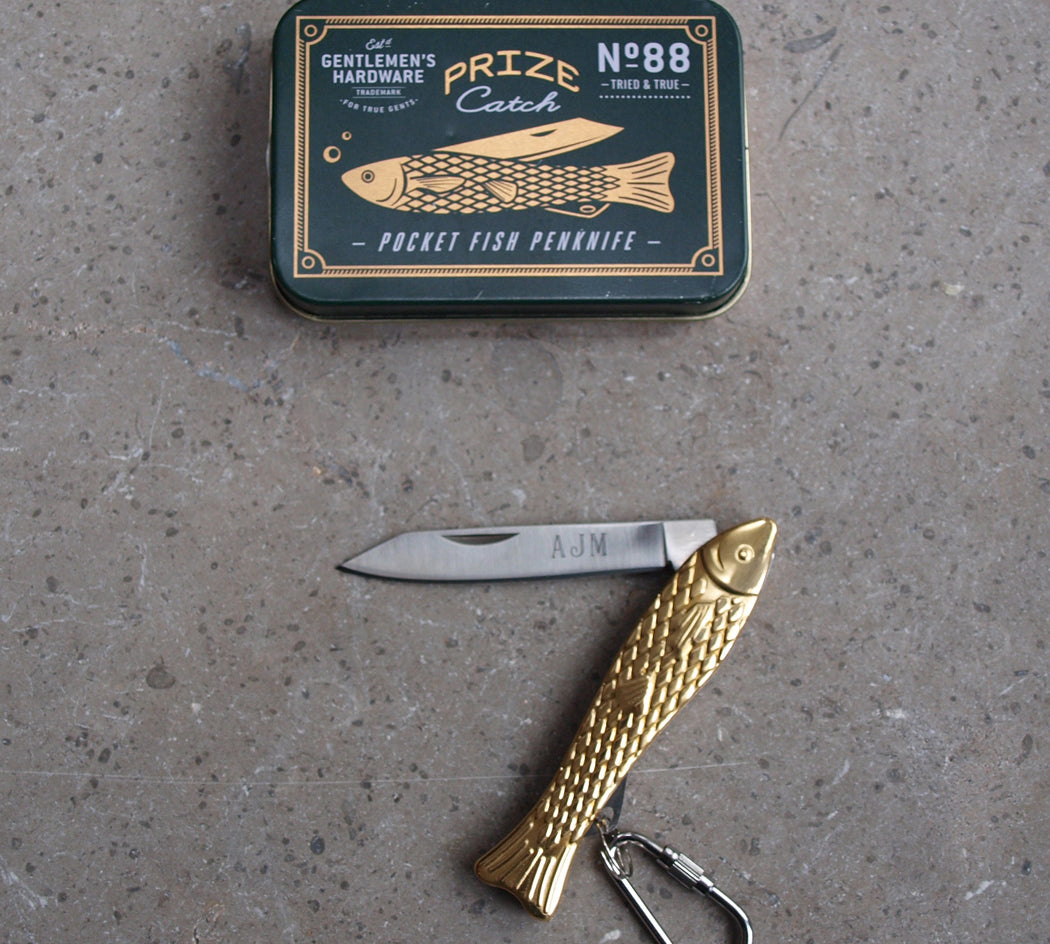 Gold fish-shaped pocket knife with a black tin labeled 'Gentlemen's Hardware' on a gray surface.