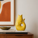 Yellow fish-shaped ceramic vase on books with a framed abstract painting in the background.
