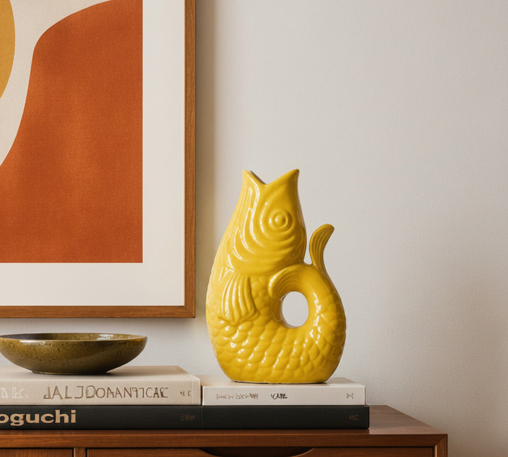 Yellow fish-shaped ceramic vase on books with a framed abstract painting in the background.