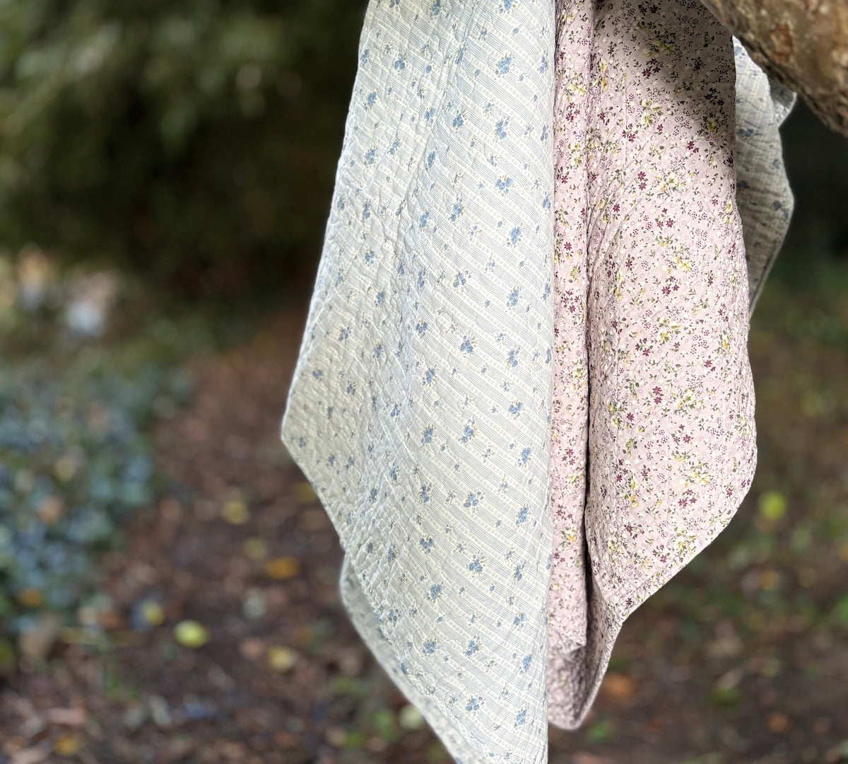 Two soft fabric floral throws hanging outdoors from a tree branch. One cloth is a light cream colour with a small blue floral pattern, and the other is a pale lilac with a dense small floral print.