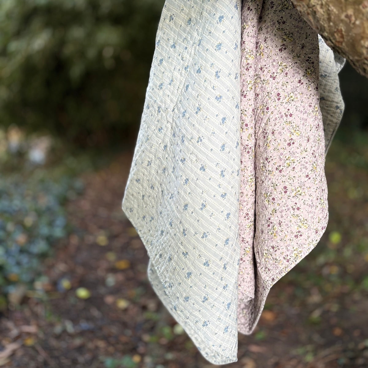 Two soft fabric floral throws hanging outdoors from a tree branch. One cloth is a light cream colour with a small blue floral pattern, and the other is a pale lilac with a dense small floral print.
