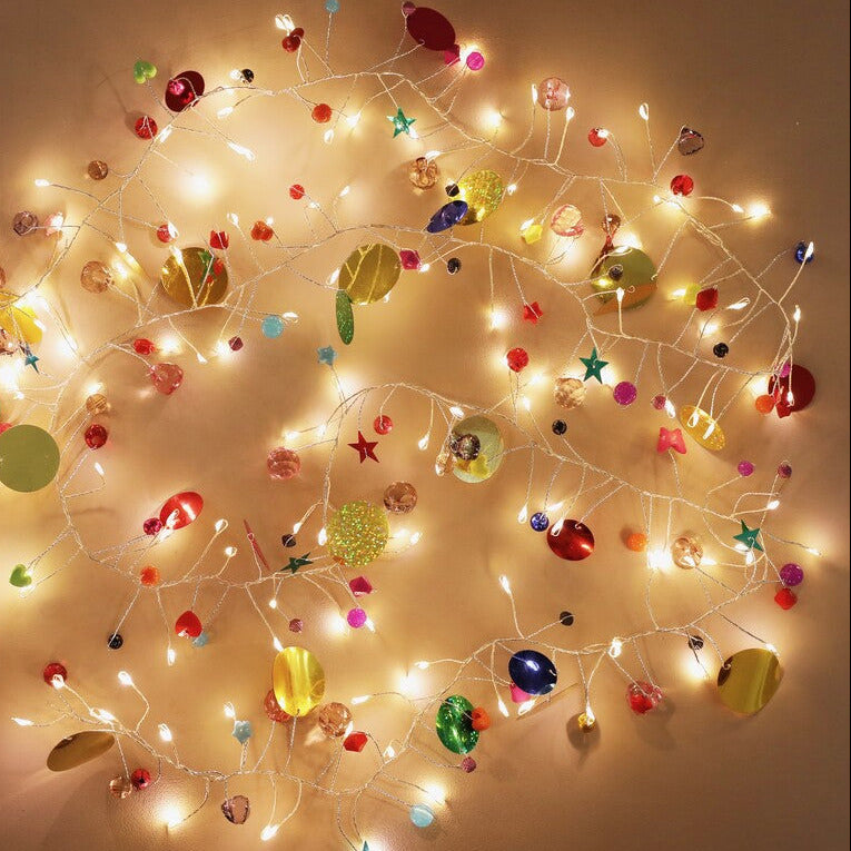 Warm white string fairy lights decorated with colorful reflective ornaments, including planets, stars, and round baubles, arranged in a circular pattern against a plain beige wall.
