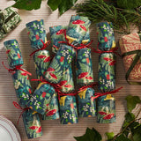 A set of Christmas-themed paper cracker gifts wrapped in a festive forest pattern with red mushrooms, green foliage, and red ribbons, arranged on a textured beige tablecloth with holiday greenery and other wrapped presents.