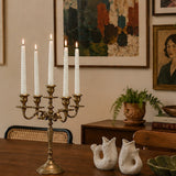 gold candlestick holder with white candles on a wooden table in a room with framed artwork on the wall.