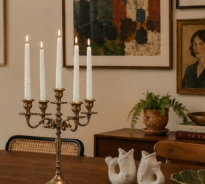 gold candlestick holder with white candles on a wooden table in a room with framed artwork on the wall.