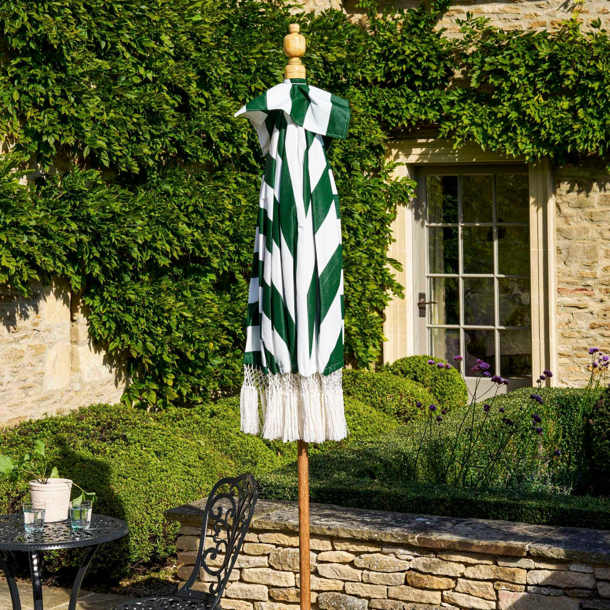 A closed patio umbrella with bold green and white diagonal stripes and a white fringed hem, displayed on a wooden stand in an outdoor garden patio setting with stone walls and a window.