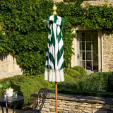 A closed patio umbrella with bold green and white diagonal stripes and a white fringed hem, displayed on a wooden stand in an outdoor garden patio setting with stone walls and a window.