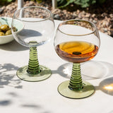 Two wine glasses on a table with one containing a drink, set against a natural outdoor background.