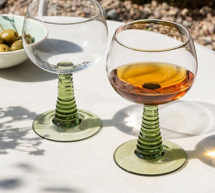 Two wine glasses on a table with one containing a drink, set against a natural outdoor background.