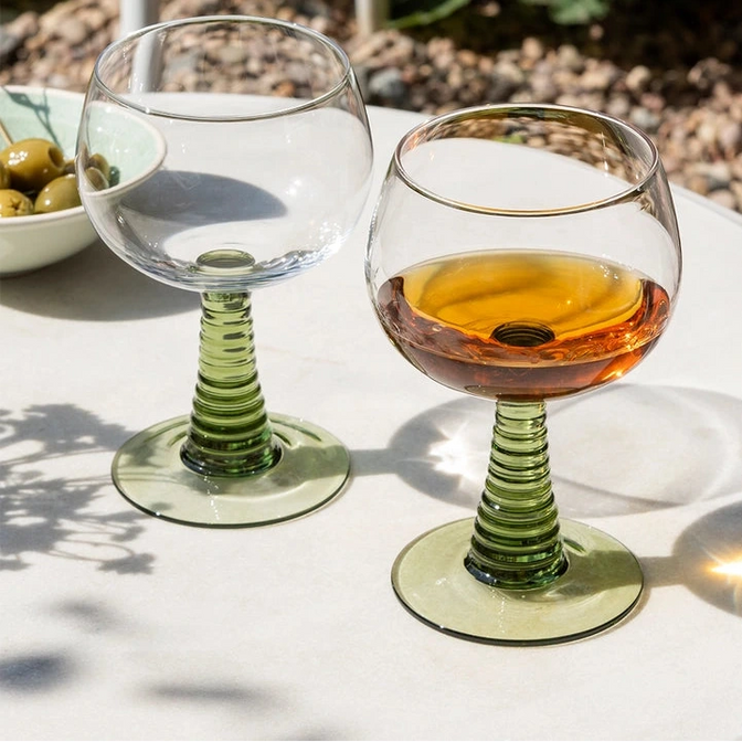 Two wine glasses on a table with one containing a drink, set against a natural outdoor background.