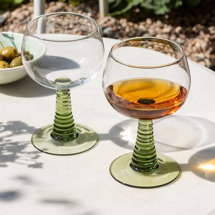 Two wine glasses on a table with one containing a drink, set against a natural outdoor background.