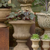 A set of stone garden planters displayed on a rustic wooden shelf, including a large textured beige urn planter with ornate foliage, a larger gray carved stone planter, and two small matching beige planter pots with succulents.