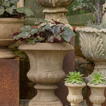 A set of stone garden planters displayed on a rustic wooden shelf, including a large textured beige urn planter with ornate foliage, a larger gray carved stone planter, and two small matching beige planter pots with succulents.