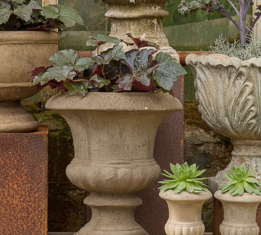 A set of stone garden planters displayed on a rustic wooden shelf, including a large textured beige urn planter with ornate foliage, a larger gray carved stone planter, and two small matching beige planter pots with succulents.