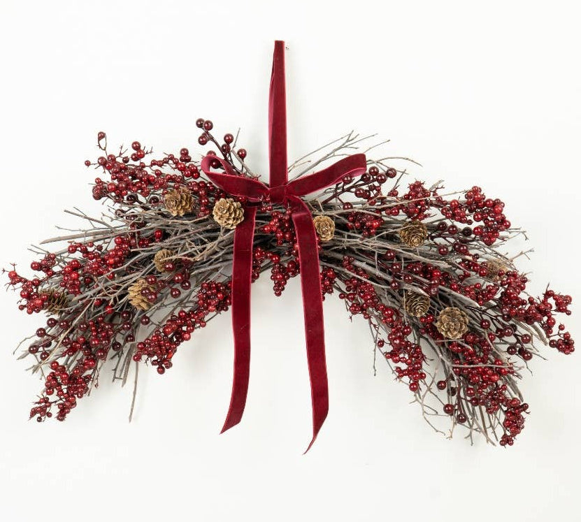 A festive Christmas wreath made of gray twig branches, covered in clusters of small red berries, with a large deep red velvet ribbon bow at the top and a few small pinecones, shown hanging against a plain white wall.