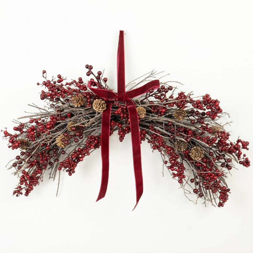 A festive Christmas wreath made of gray twig branches, covered in clusters of small red berries, with a large deep red velvet ribbon bow at the top and a few small pinecones, shown hanging against a plain white wall.