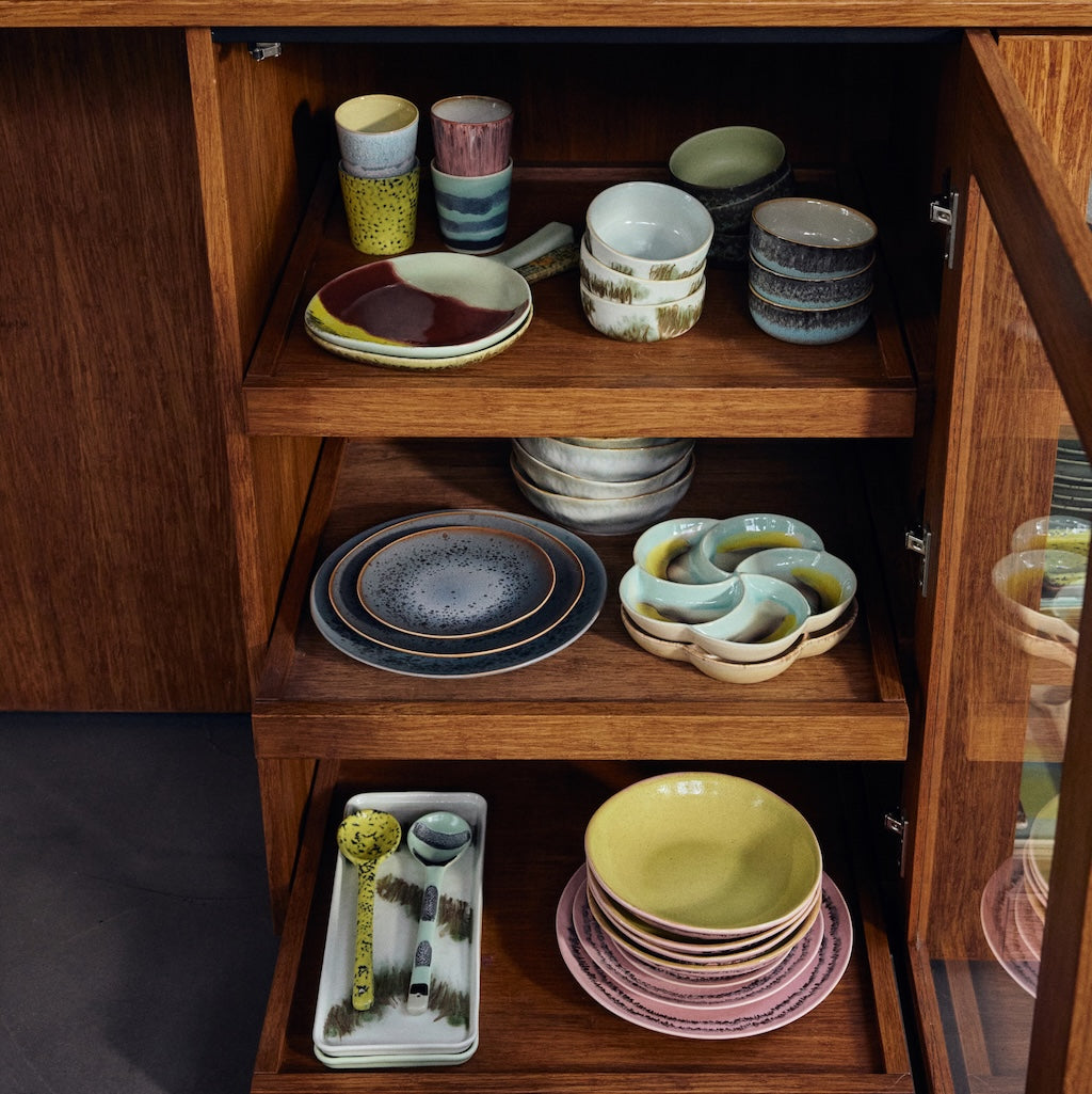A wooden cabinet with open shelves displaying a collection of hand-painted ceramic tableware, including bowls, plates, mugs, and serving dishes in various earthy colors.