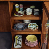 A wooden cabinet with open shelves displaying a collection of hand-painted ceramic tableware, including bowls, plates, mugs, and serving dishes in various earthy colors.