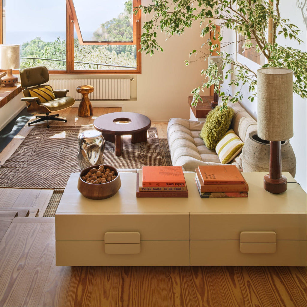 A modern living room interior with a cream sofa, striped green and cream pillows, a low round wooden coffee table, a cream storage cabinet in the foreground, and a large potted tree. A large window on the left offers a view of trees and water.