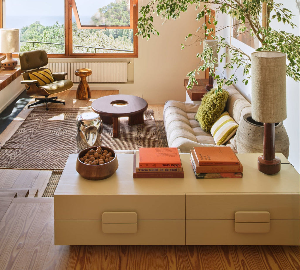 A modern living room interior with a cream sofa, striped green and cream pillows, a low round wooden coffee table, a cream storage cabinet in the foreground, and a large potted tree. A large window on the left offers a view of trees and water.