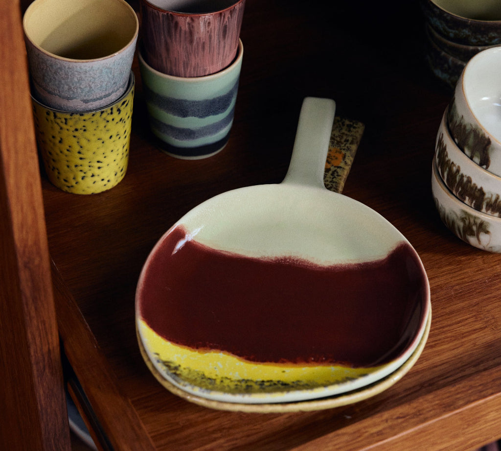 A ceramic serving bowl with a dark red and yellow marbled glaze sits on a wooden shelf, surrounded by other colorful stoneware cups and dishes.