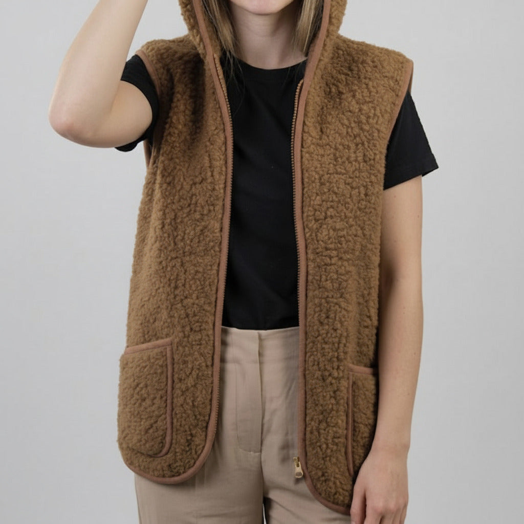 A person wearing a brown sherpa fleece zip-up hooded vest over a black short-sleeve top and beige pants, photographed against a light gray studio background.