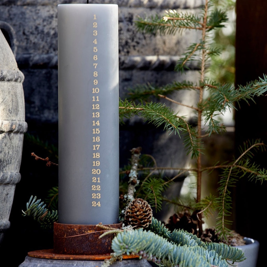 Gray advent candle in rustic candle holder, surrounded by greenery and a stone wall.