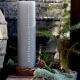Gray advent candle in rustic candle holder, surrounded by greenery and a stone wall.