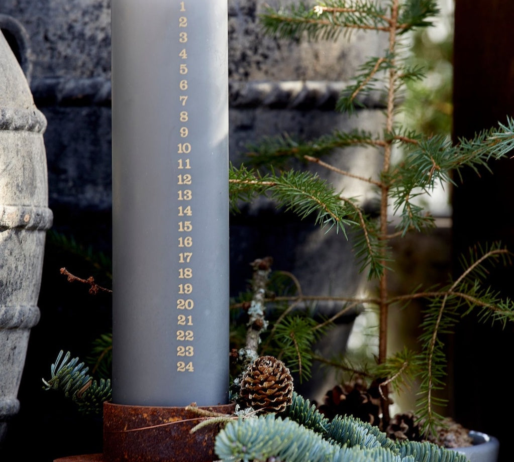 Gray advent candle in rustic candle holder, surrounded by greenery and a stone wall.