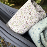 A close-up of a floral quilted pillow resting on a gray fabric hammock, with a rolled light blue patterned quilt underneath in an outdoor setting.