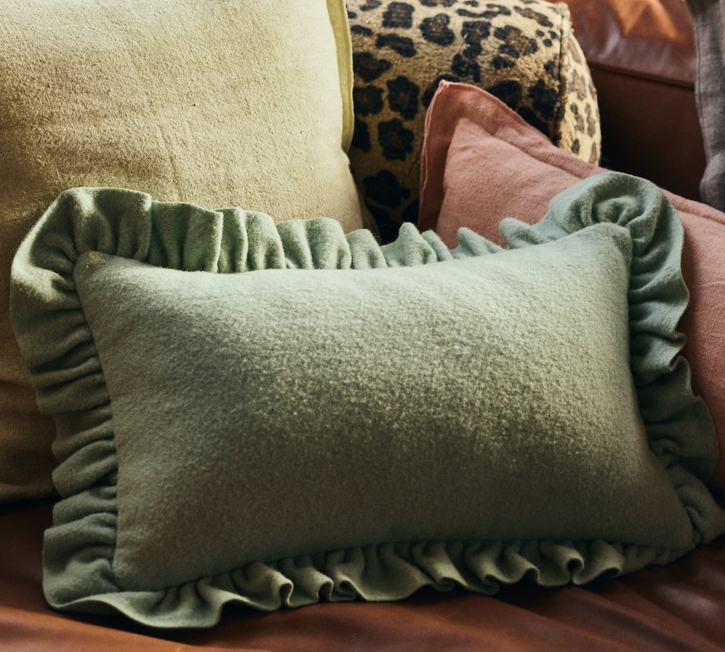 An mint green rectangular cushion with ruffled trim, styled on a brown leather sofa alongside other decorative pillows.