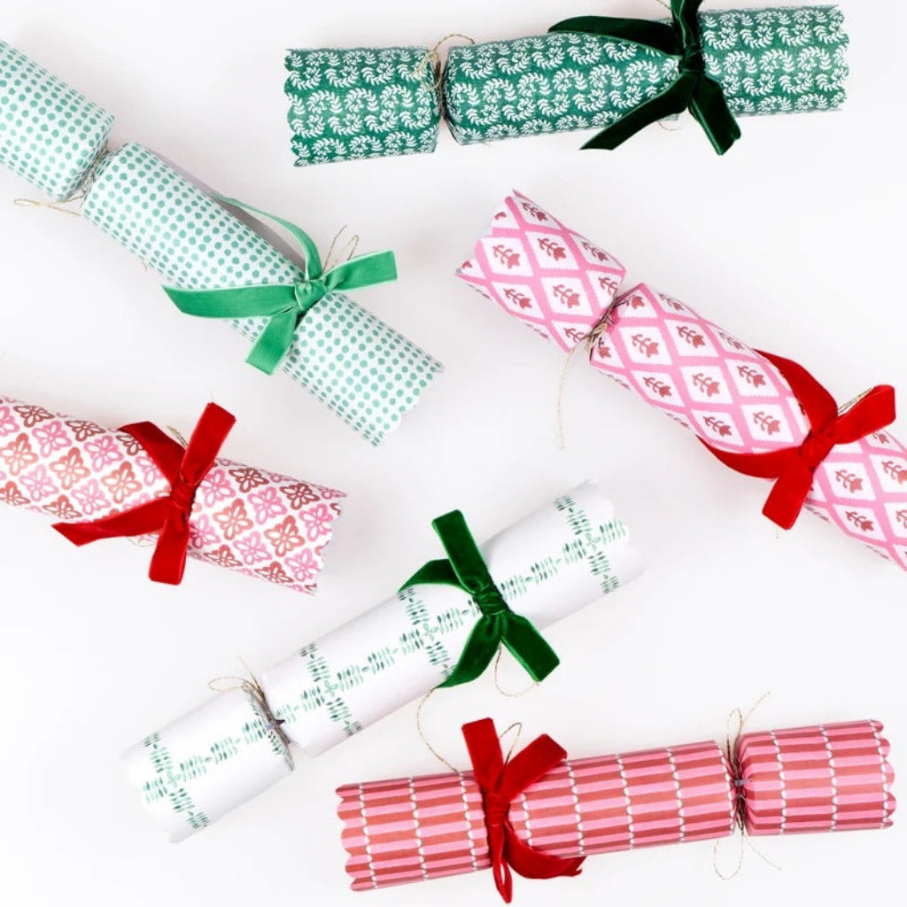Six Christmas cracker ornaments wrapped in festive patterned paper and tied with green and red ribbons, arranged on a clean white background.