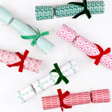 Six Christmas cracker ornaments wrapped in festive patterned paper and tied with green and red ribbons, arranged on a clean white background.