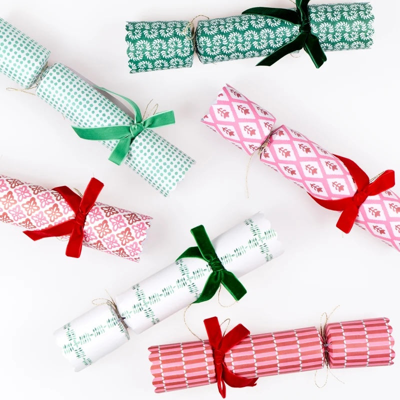Six Christmas cracker ornaments wrapped in festive patterned paper and tied with green and red ribbons, arranged on a clean white background.